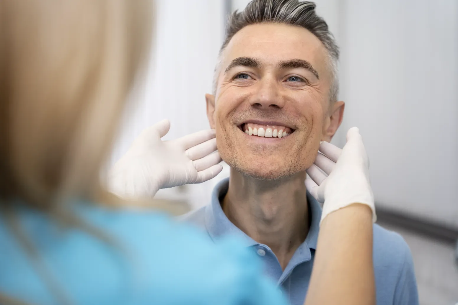 A man with gray hair smiles while a healthcare professional wearing white gloves examines his neck.