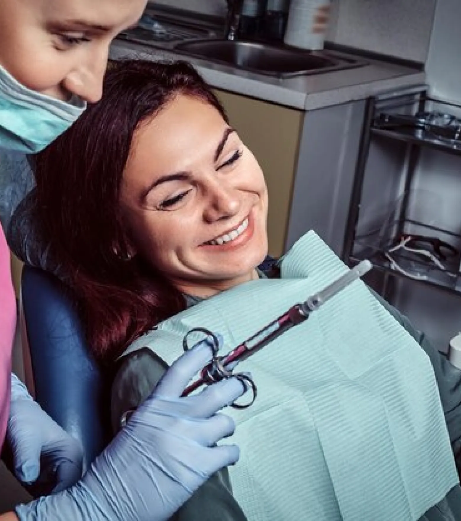 Smiling female patient in a dental chair looking at a syringe held by a masked dental professional wearing gloves.