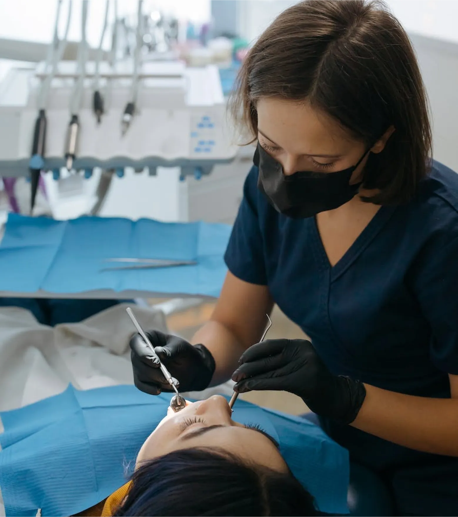 Dentist wearing black gloves and mask examining a patient's teeth using dental instruments.