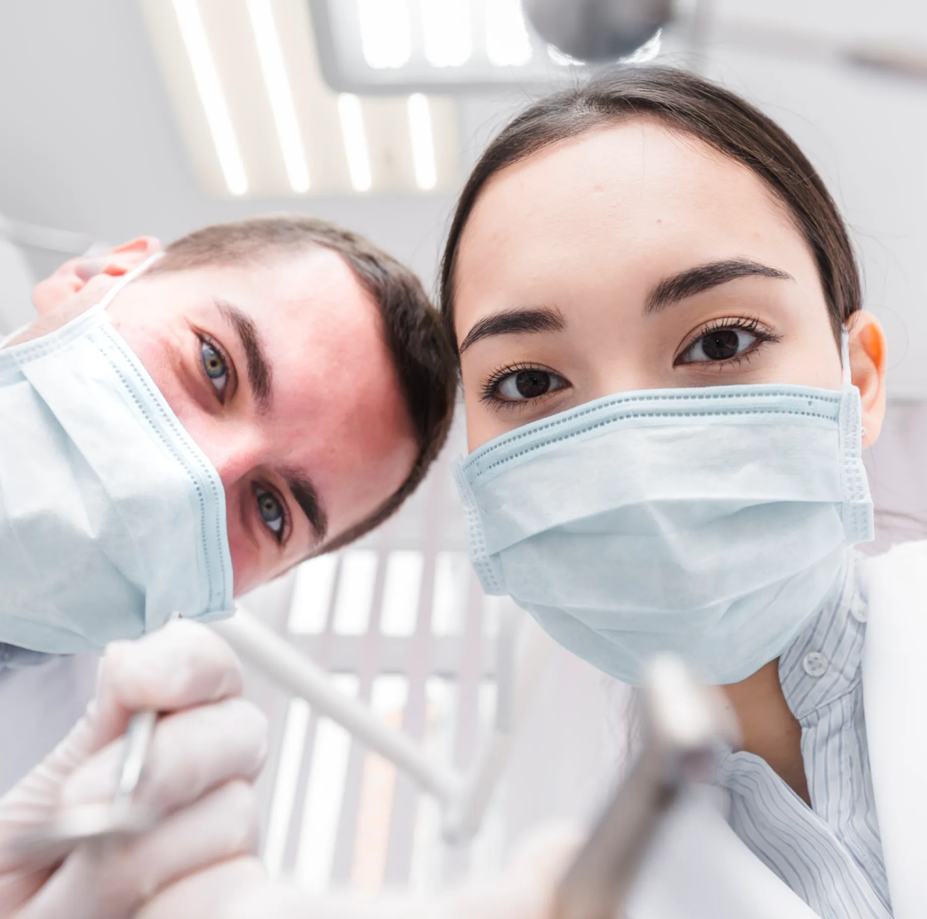 Two dentists wearing surgical masks and gloves leaning toward the camera with dental tools.