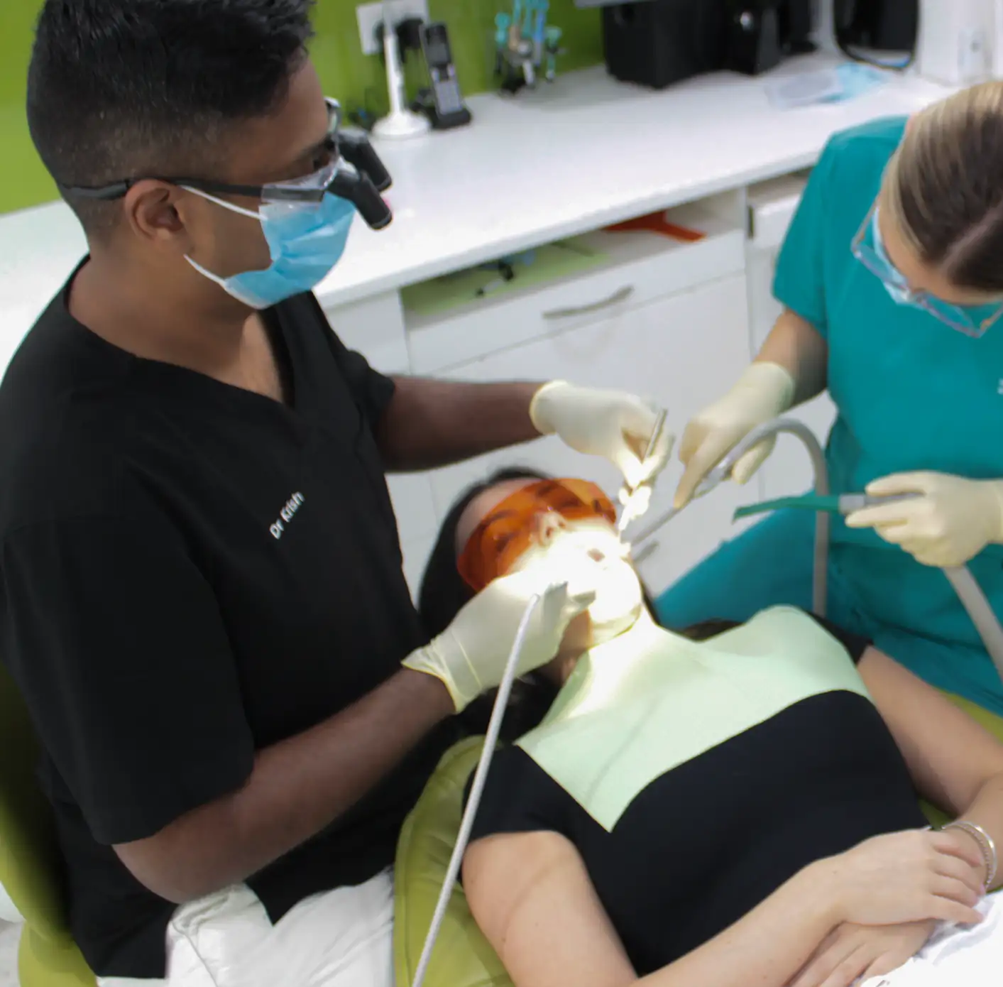Dentist and dental assistant wearing masks and gloves treating a patient reclining in a dental chair.