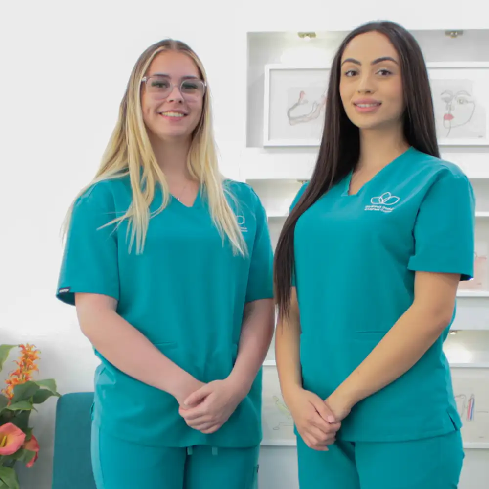 Two female healthcare professionals wearing teal scrubs standing and smiling in a clinical setting.