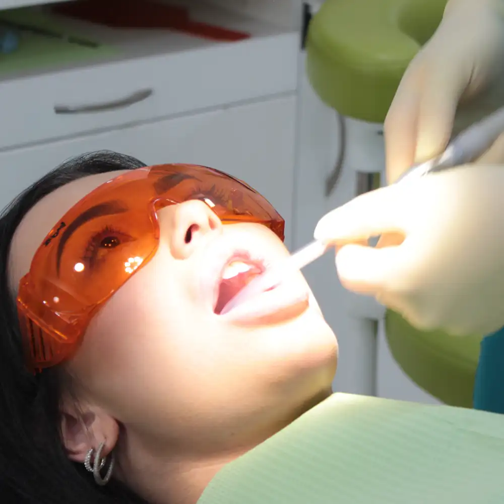 Patient wearing orange protective glasses receiving dental treatment from a gloved dentist using a dental tool.