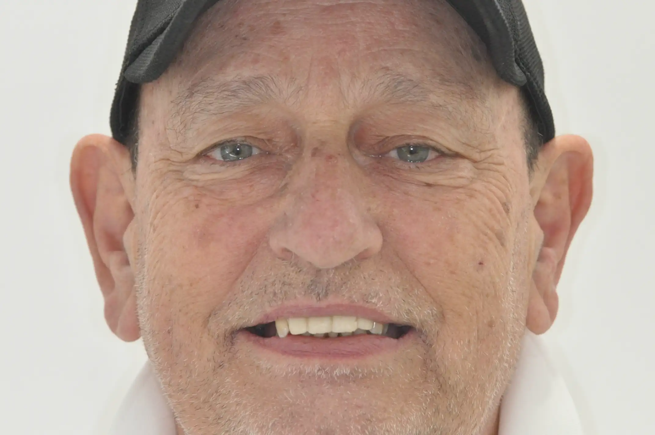 Close-up of a smiling older man with blue eyes, light stubble, wearing a black cap and white shirt against a plain background.