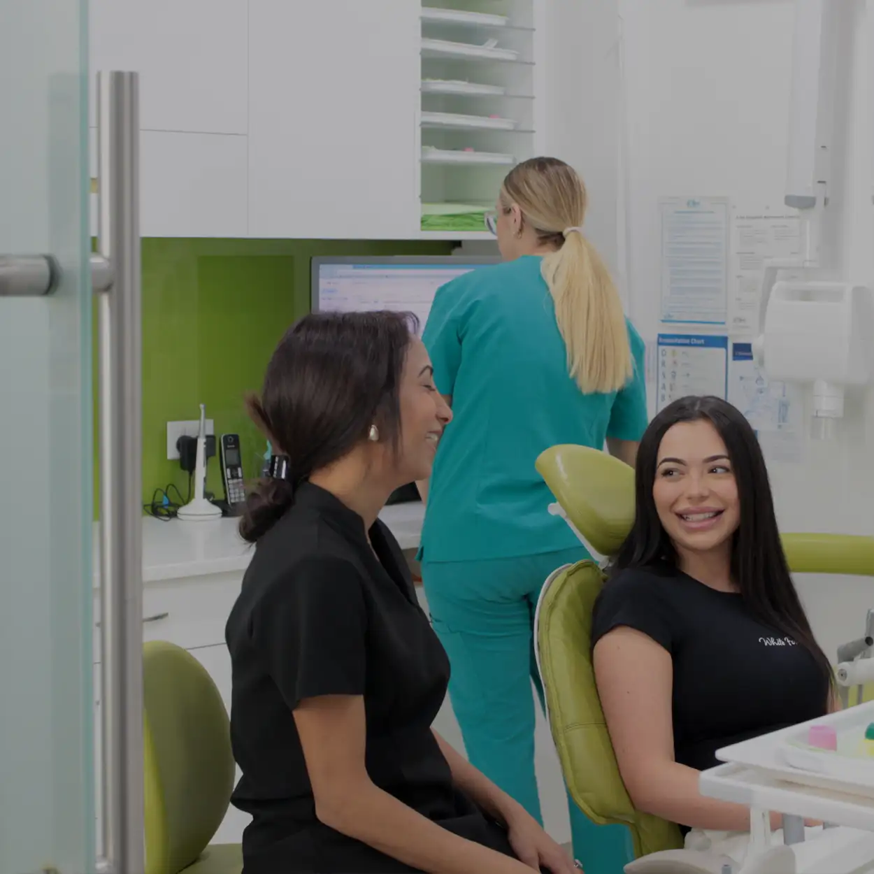 Two women, one in black scrub and the other in black t-shirt sitting and smiling at each other in a dental office, with a dental professional in teal scrubs working at a computer in the background.