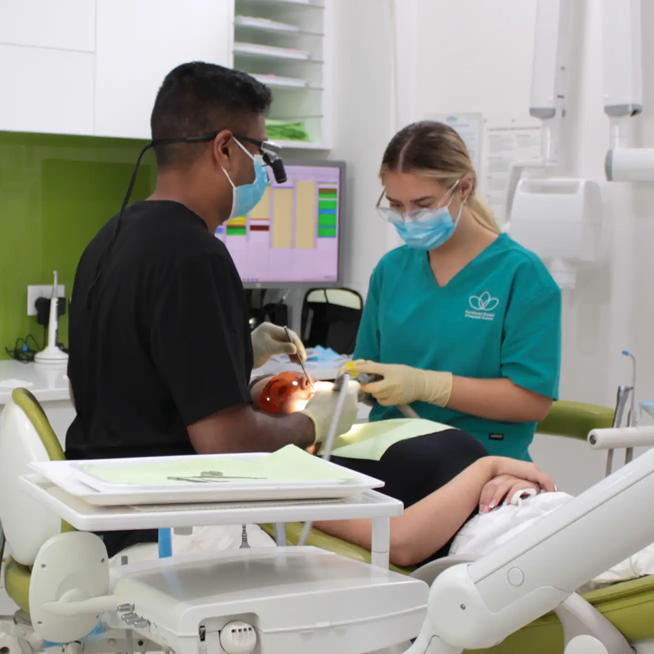 Two dental professionals wearing face masks and gloves treating a patient lying on a dental chair in a clinic.