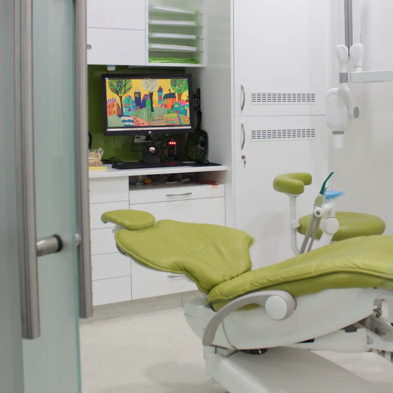 Modern dental clinic room with a green dental chair and a computer monitor displaying colorful artwork.