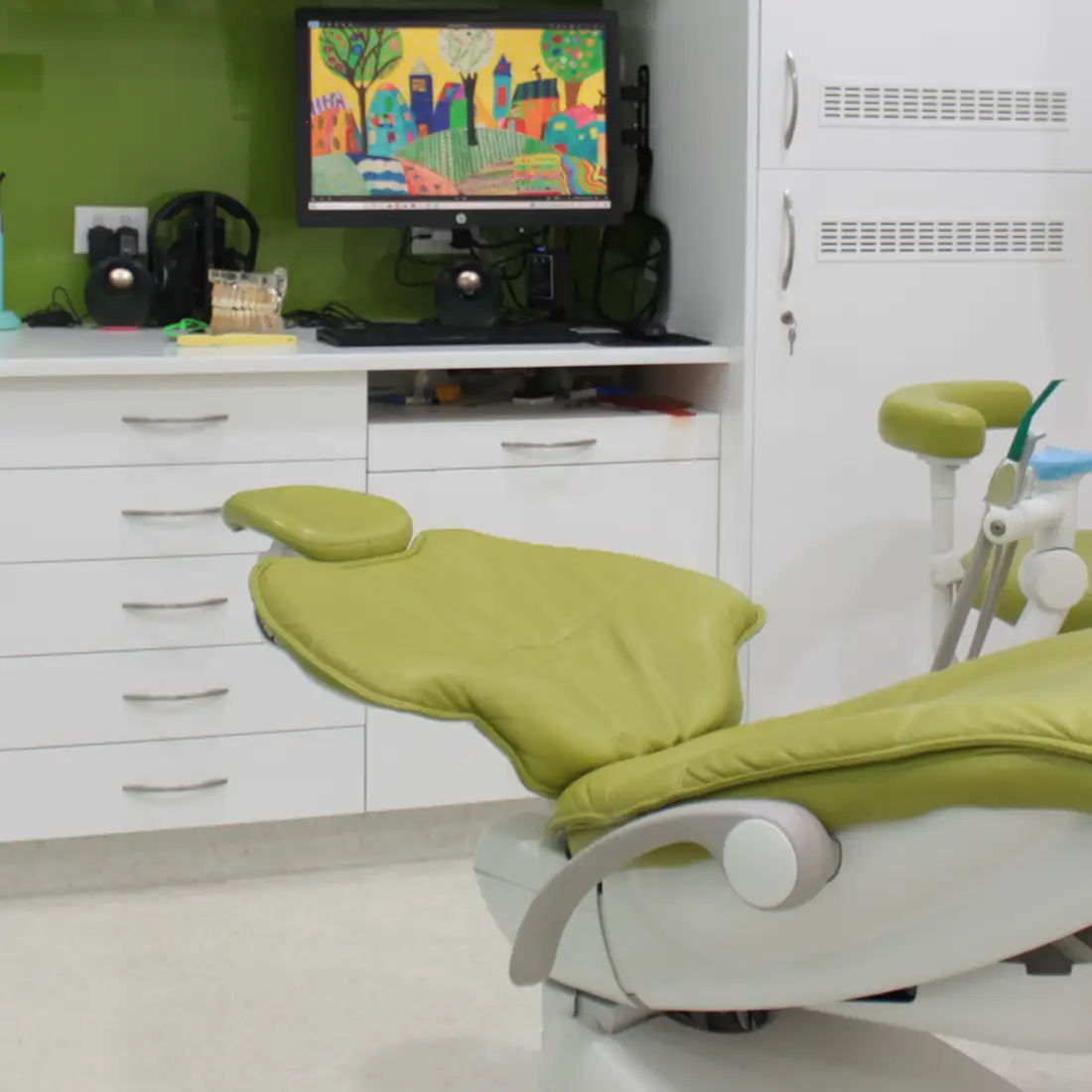 Green dental chair in a modern dentist's office with a computer monitor displaying colorful artwork in the background.