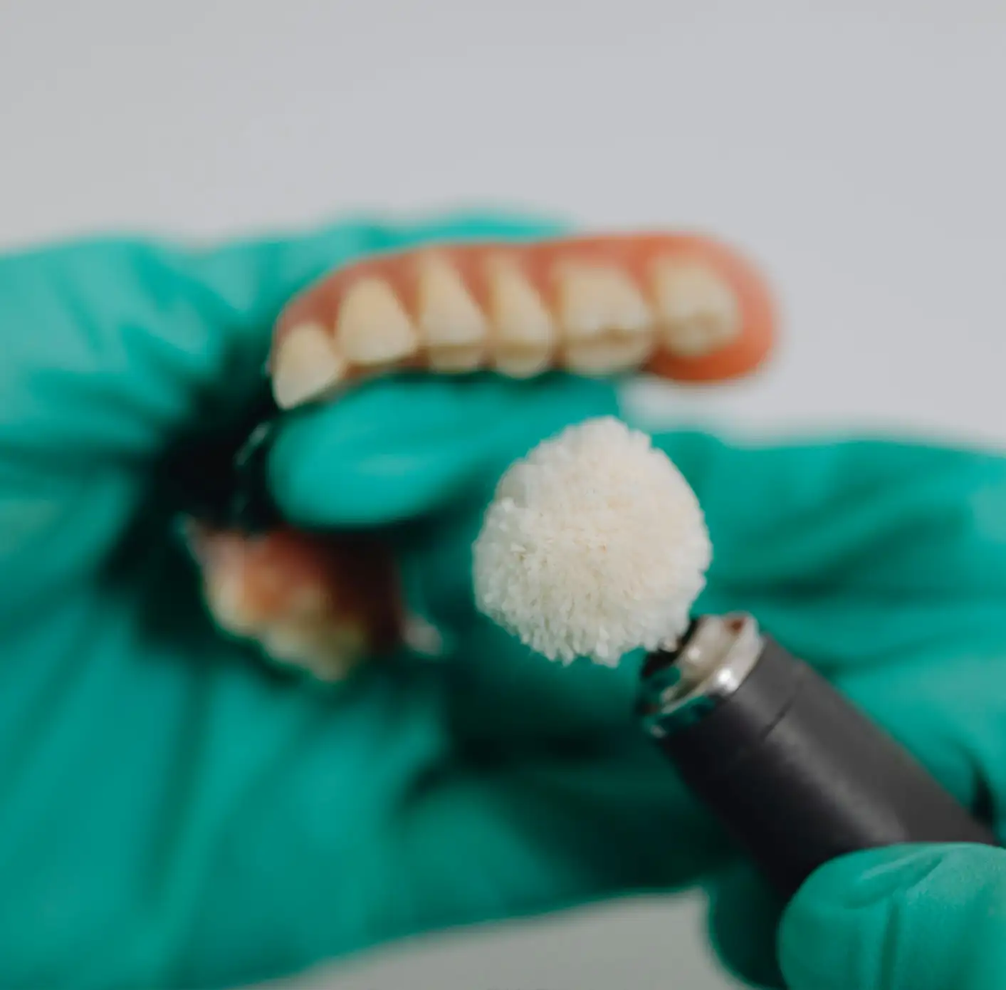 Gloved hands holding a denture and a dental polishing tool with a white buffing wheel.