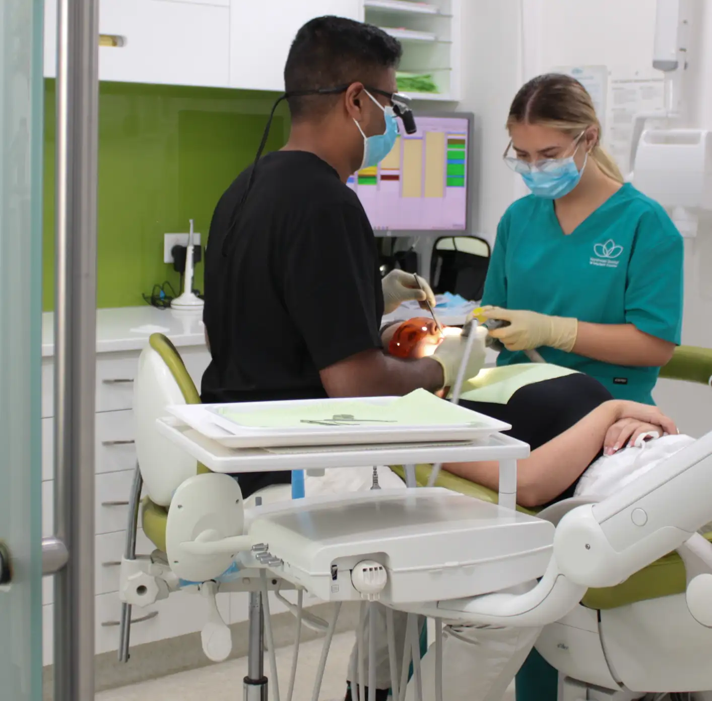 Dentist wearing magnifying glasses and a mask treating a patient while a dental assistant in scrubs holds a suction tool in a modern dental clinic.