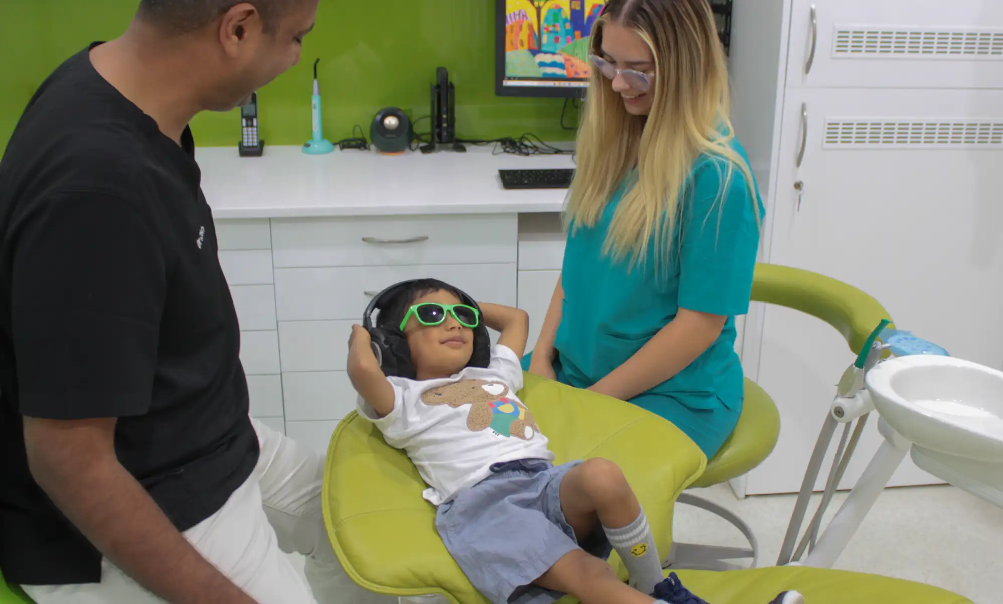 Young child wearing green sunglasses and headphones reclining in a dental chair, smiling man and woman in scrubs nearby.