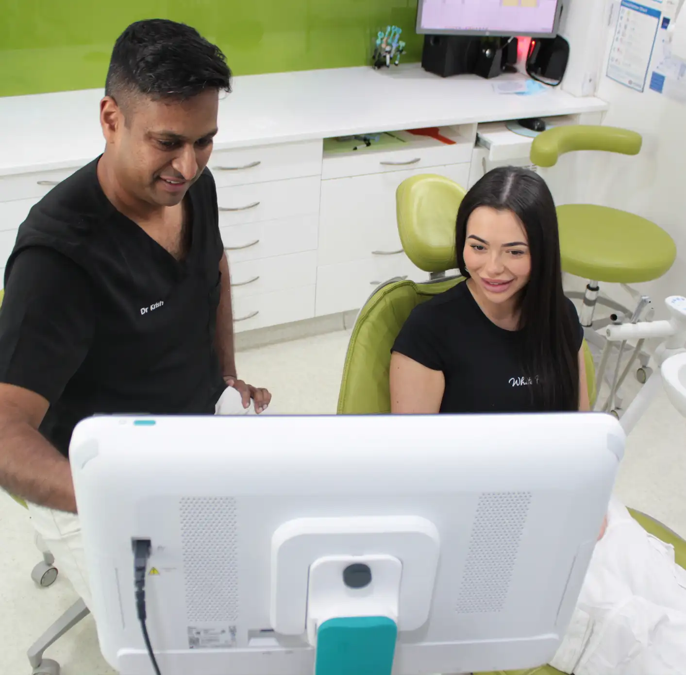 Dentist in black scrubs showing a computer screen to a female patient seated in a green dental chair in a dental clinic.