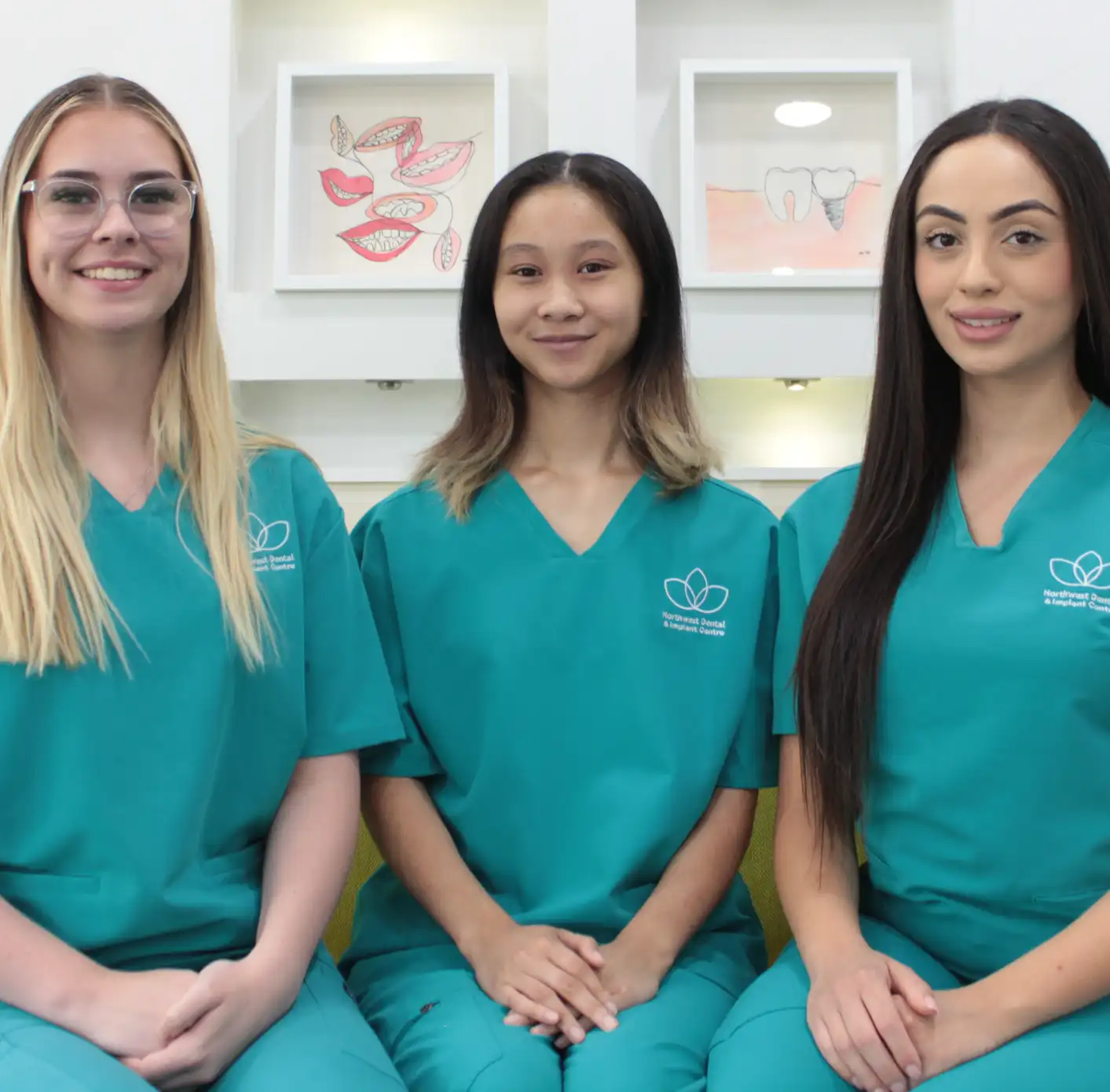 Three female dental professionals wearing teal scrubs sitting side by side smiling.