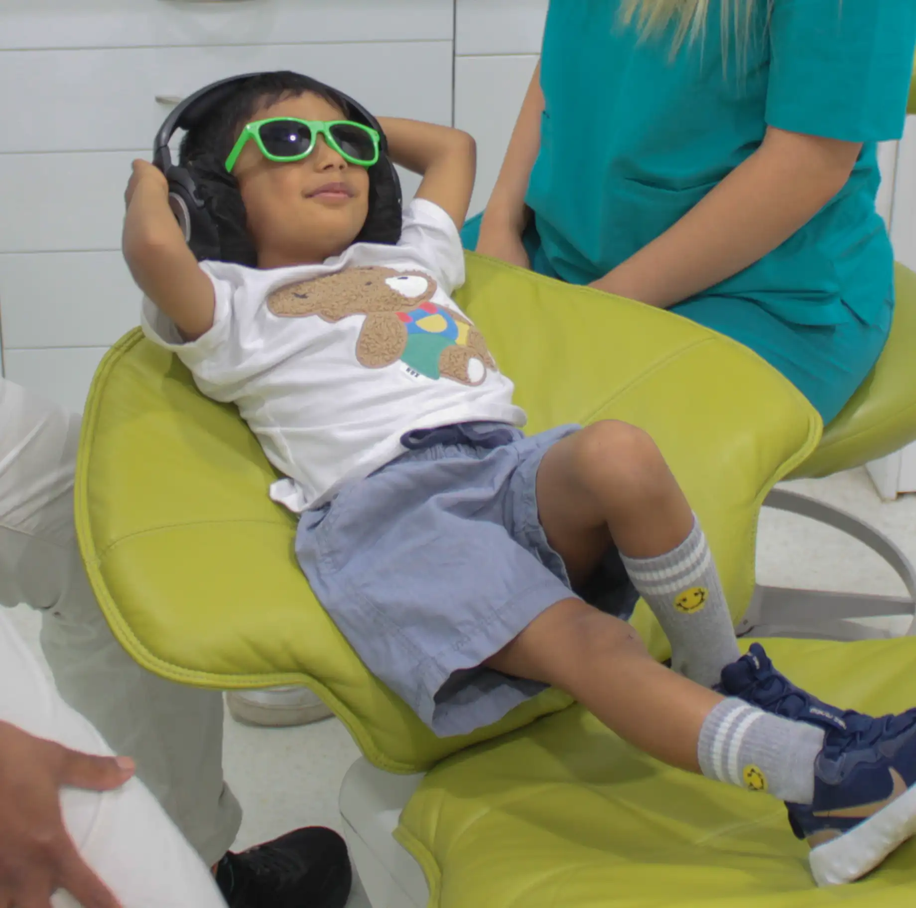 Young boy wearing green sunglasses and headphones reclining on a green dental chair with two adults nearby.