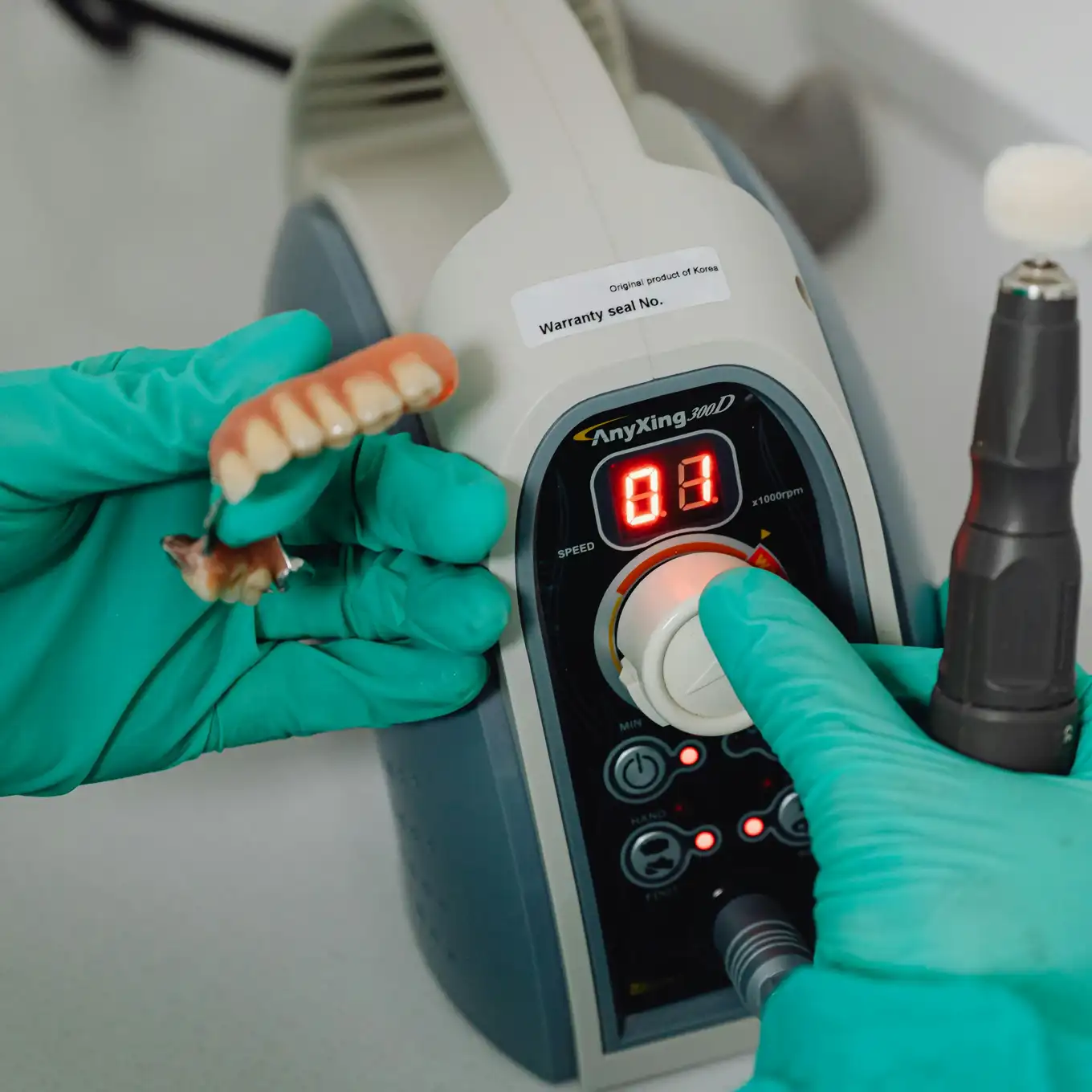 Gloved hands holding dentures and adjusting the speed dial on an dental device used for polishing dentures.