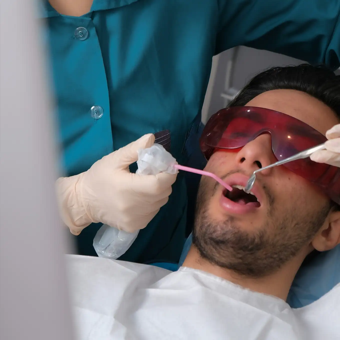 Dentist wearing gloves and a teal coat performing dental tooth extraction treatment on a patient.