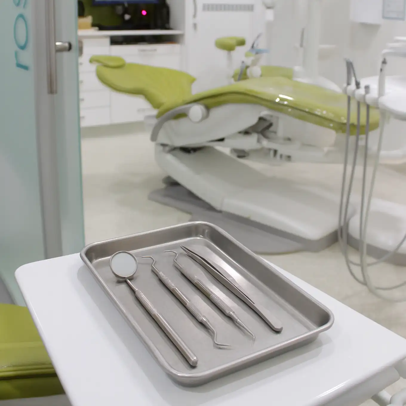 Metal tray with dental tools including a mirror, probes, and tweezers on a white table with a green dental chair in the background.