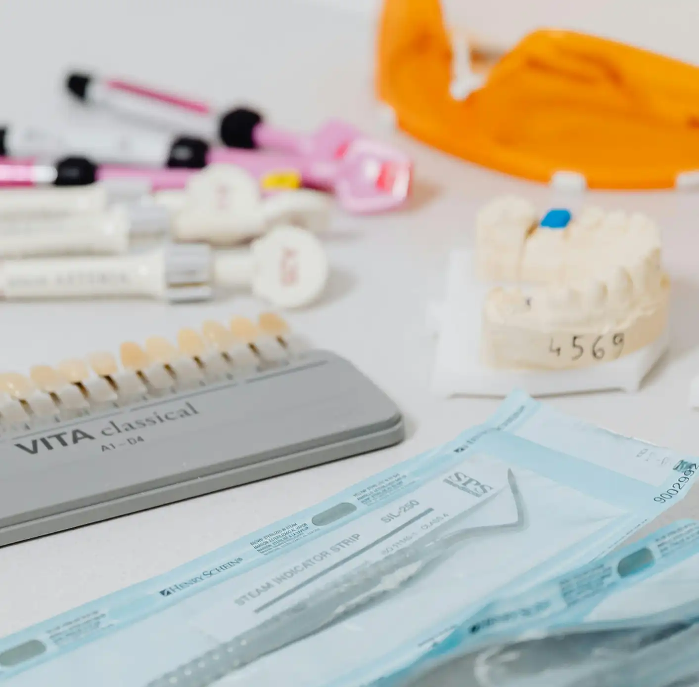 Various dental tools and materials on a white surface, including a tooth shade guide, dental impression model, and sealed dental instruments needed for dental fillings.