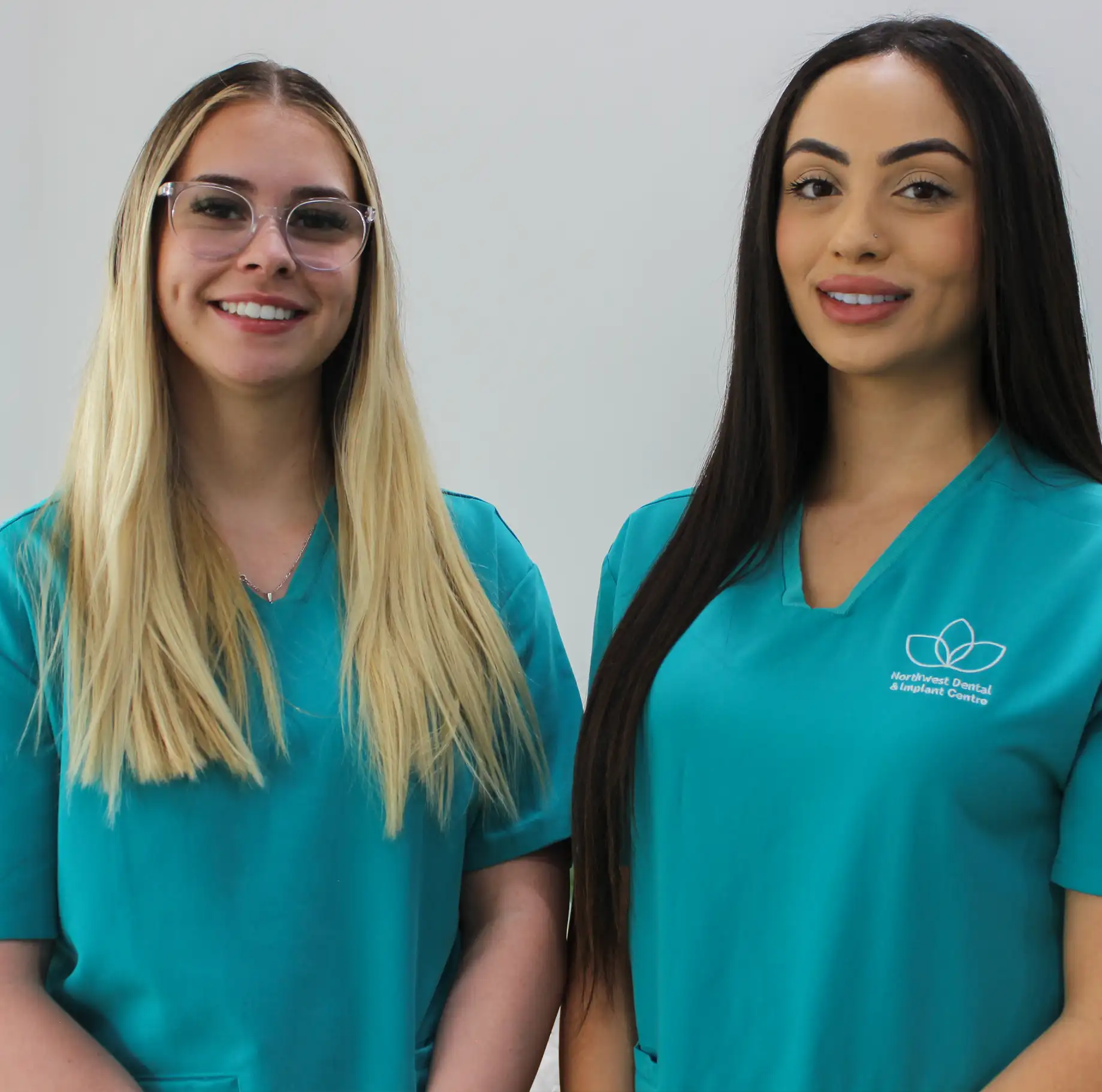 Two smiling women wearing teal medical scrubs, one with glasses and blonde hair, the other with long dark hair.