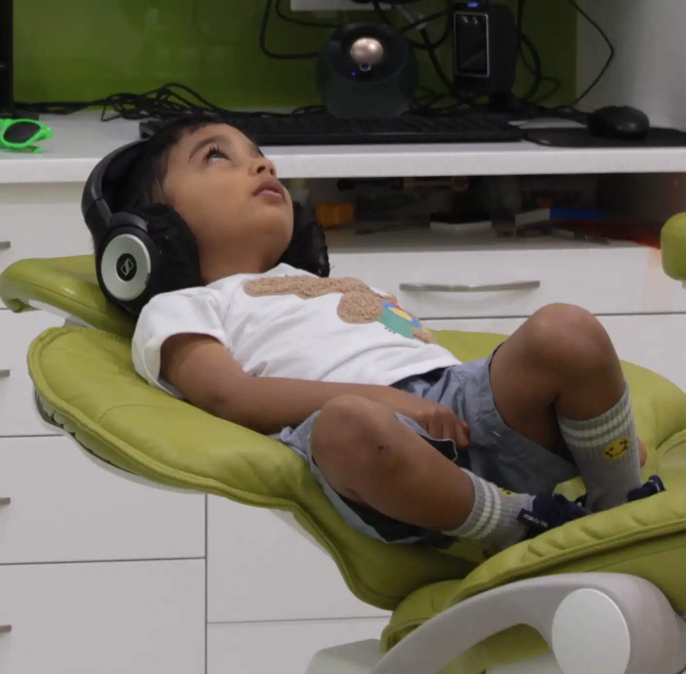 Young child lying back in a green dental chair wearing large black headphones and looking upward.