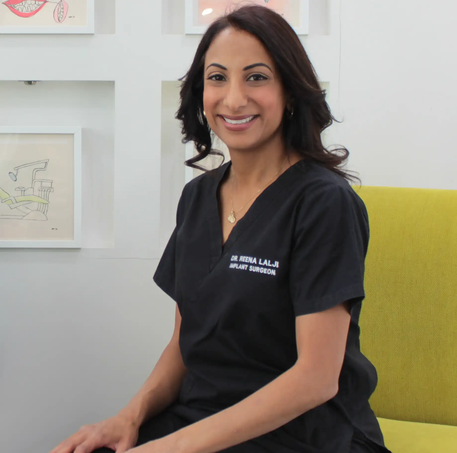 Smiling female implant surgeon in black scrubs sitting on a yellow chair in a medical office.