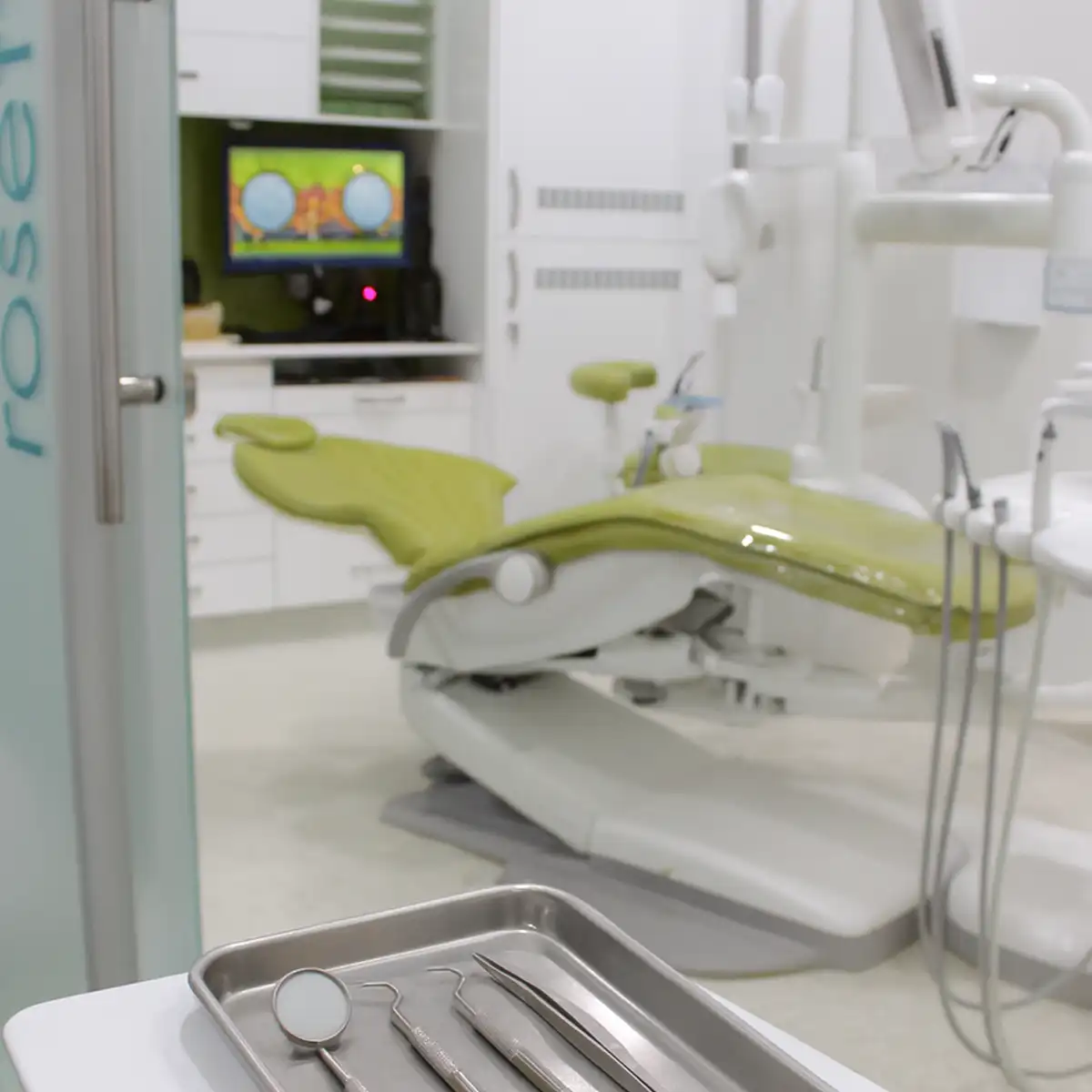 Close-up of dental tools on a tray with a green dental chair and equipment in a clean dental clinic room.