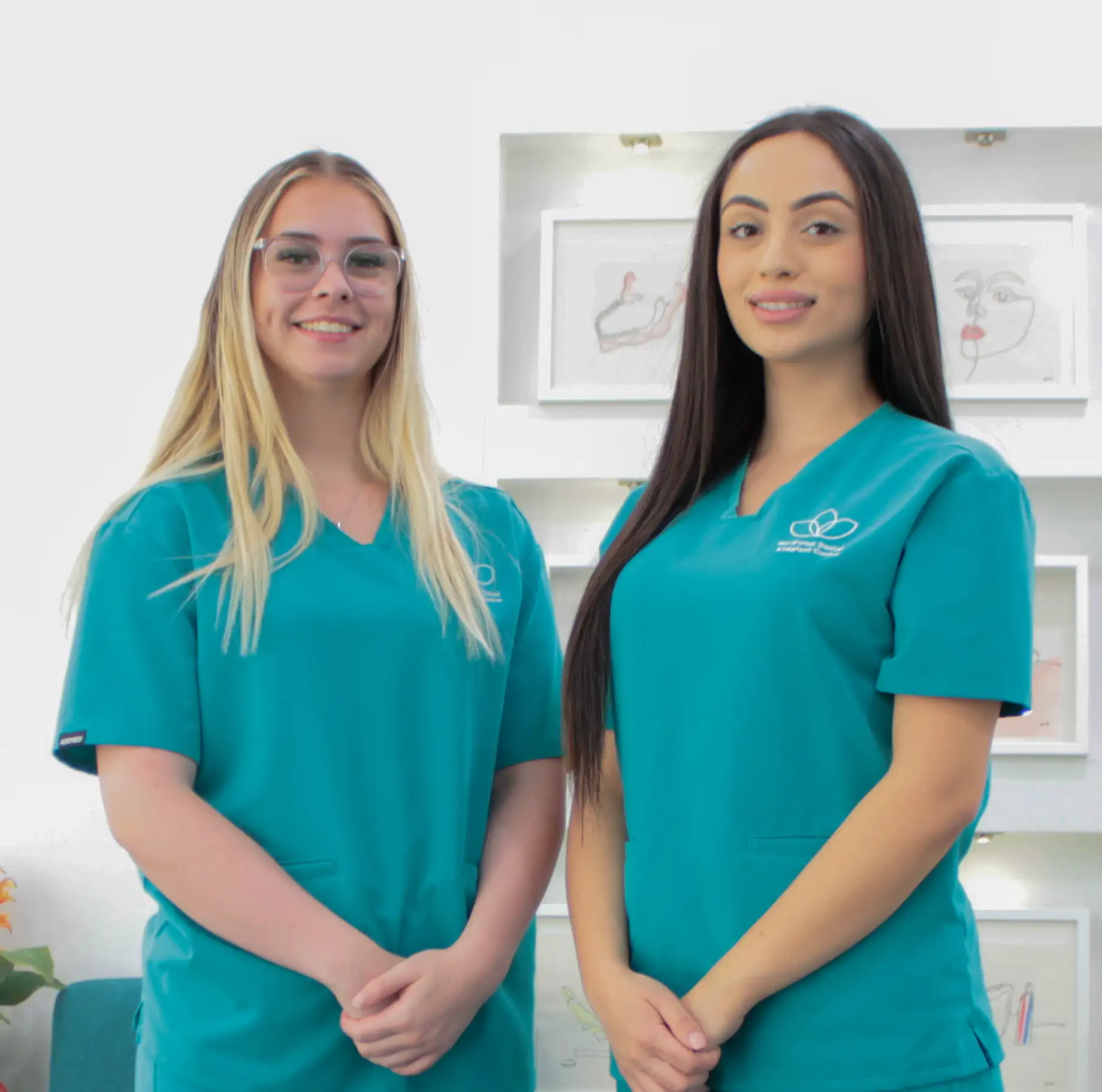 Two female healthcare professionals in teal scrubs smiling and standing indoors in a medical office.