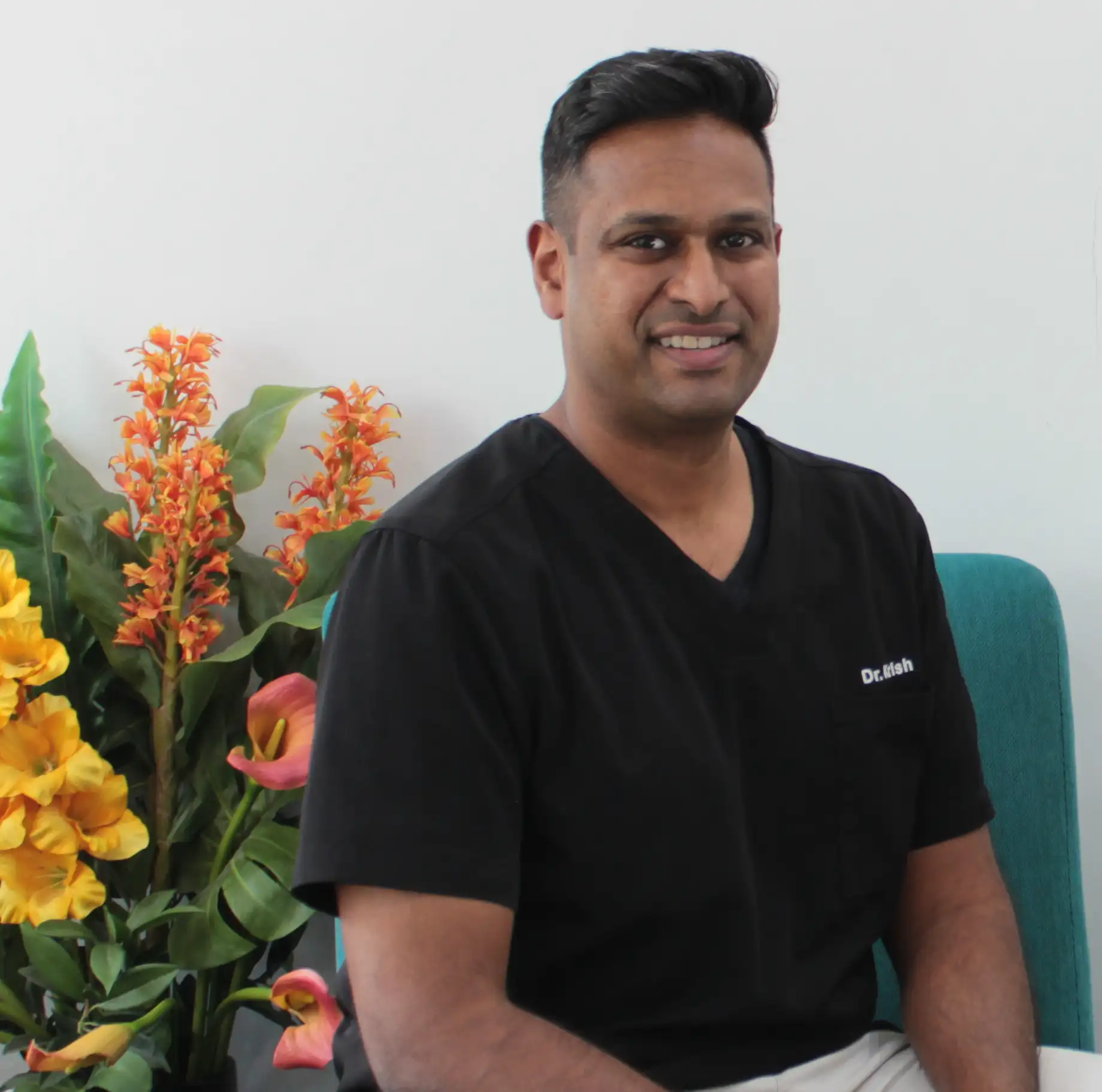 Dr. Krish  in black scrubs sitting on a teal chair with colourful flowers in the background.