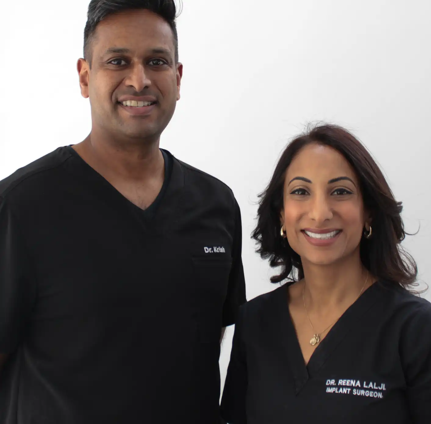 Two smiling medical professionals in black scrubs, one male with 'Dr. Krish' and one female with 'Dr. Reena Lalji Implant Surgeon' embroidered on their tops.