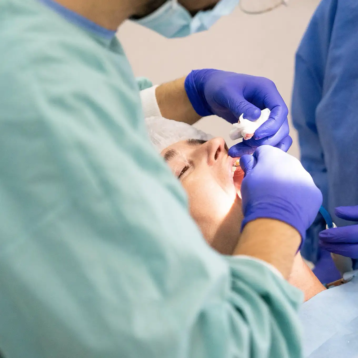 Dentist wearing blue gloves treating a patient's teeth while the patient reclines with eyes closed.