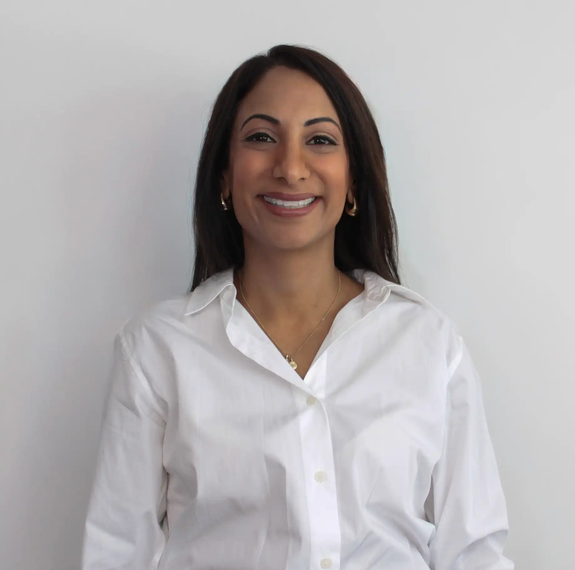 Smiling woman with long dark hair wearing a white button-up shirt and gold hoop earrings against a plain light background.