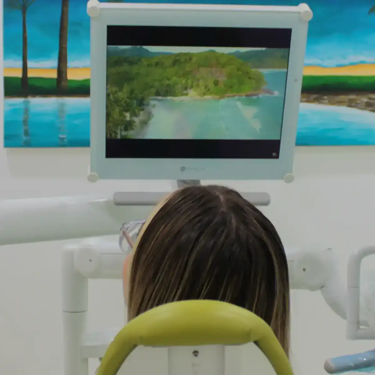 Person reclining in a dental chair watching a screen displaying a coastal landscape with water and green hills.