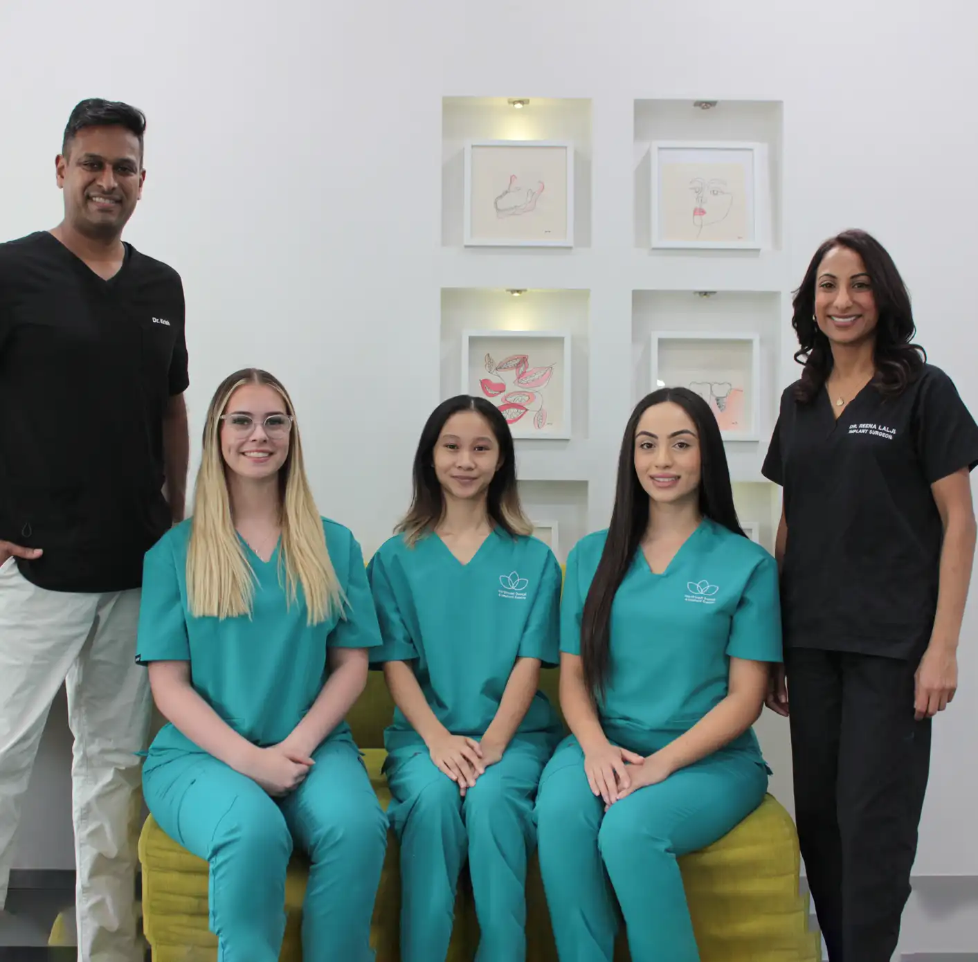 Five dental professionals, three women in teal scrubs seated and two doctors in black scrubs standing in a clinical setting with framed artwork behind them.