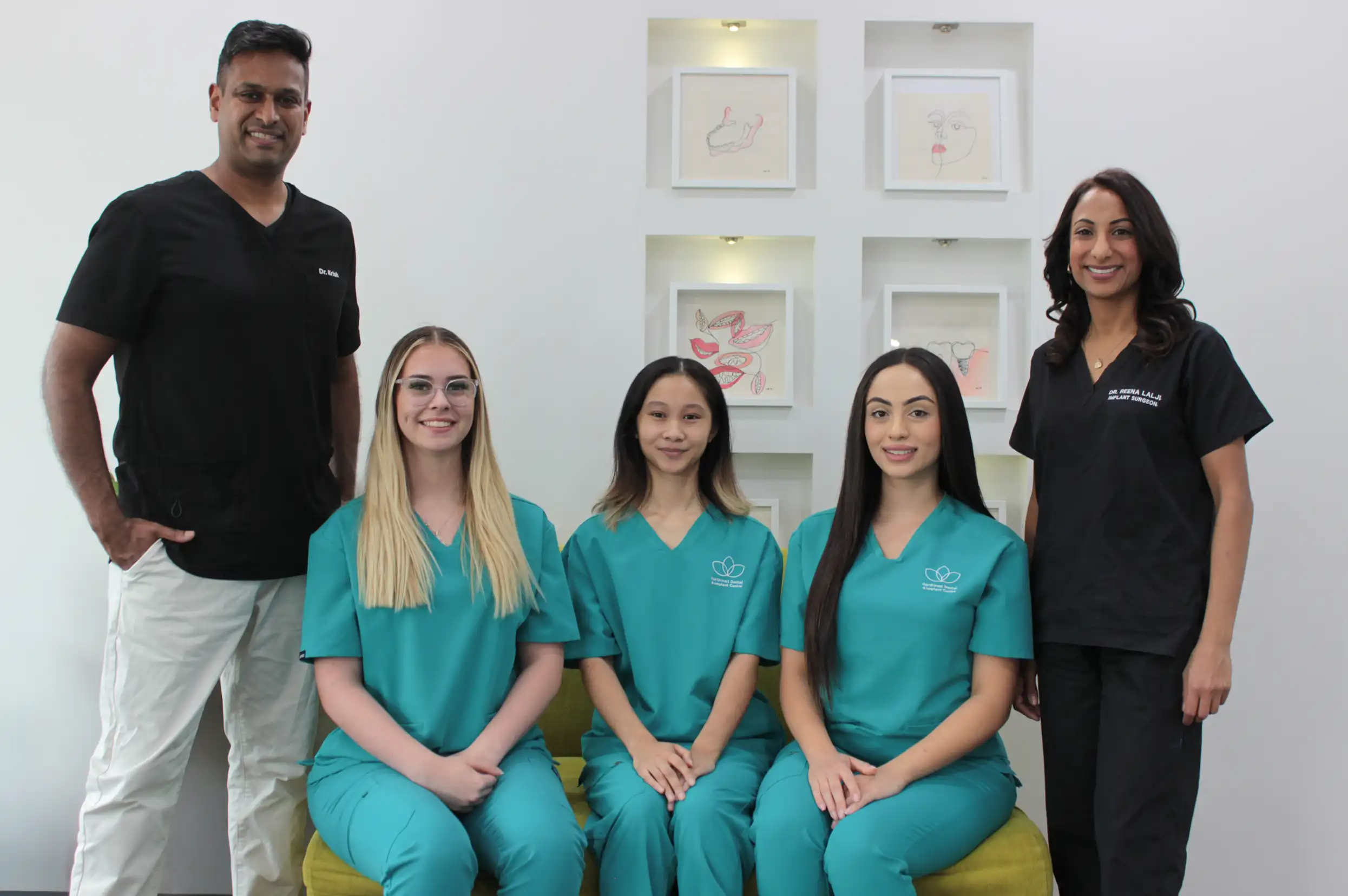Five dental professionals smiling, three women seated wearing teal scrubs and two standing wearing black scrubs, in a clinic with framed dental artwork on the wall behind them.
