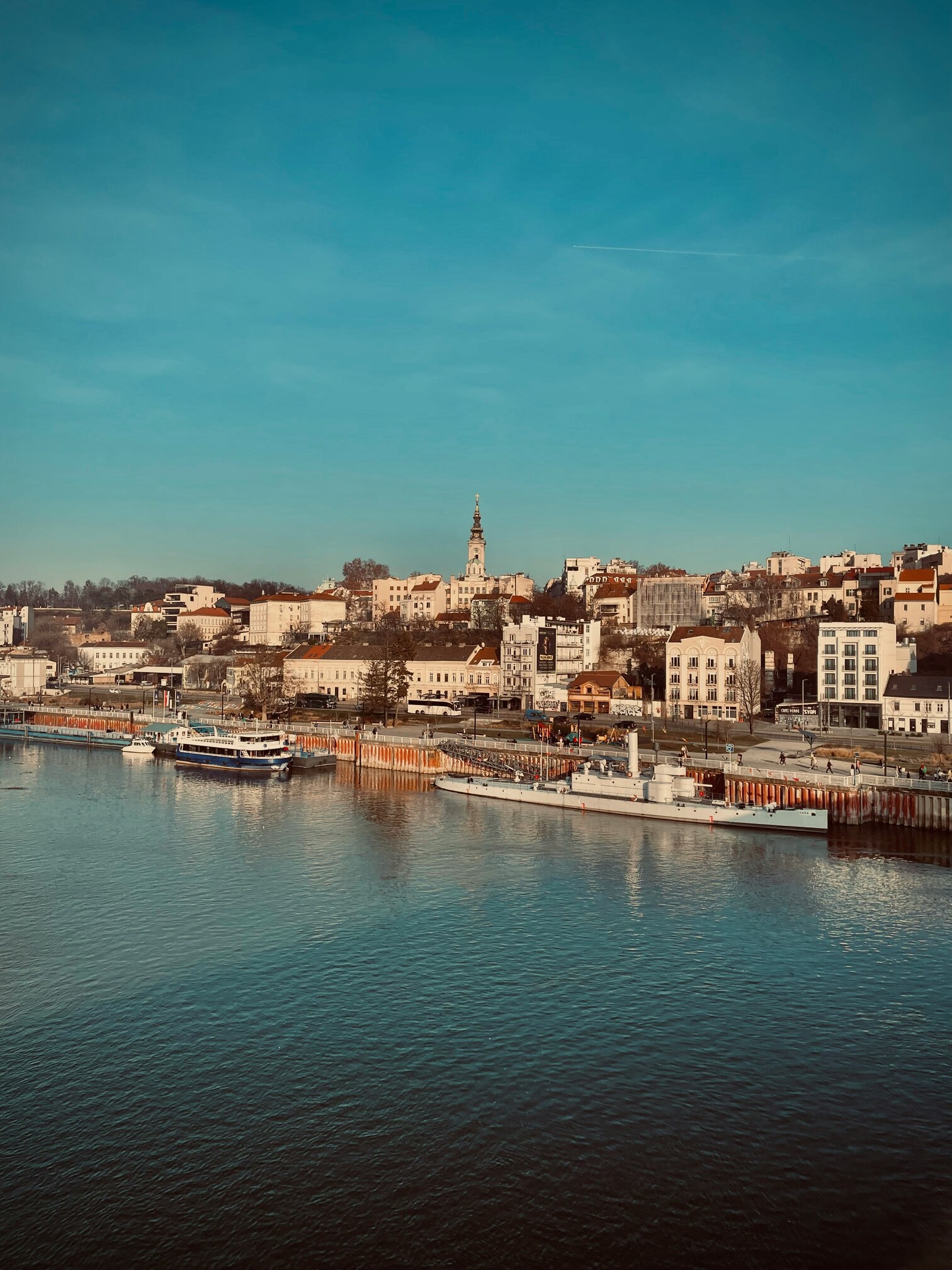 Vue sur un port avec plusieurs bateaux amarrés et une ville avec des bâtiments historiques et une église au centre sous un ciel clair.
