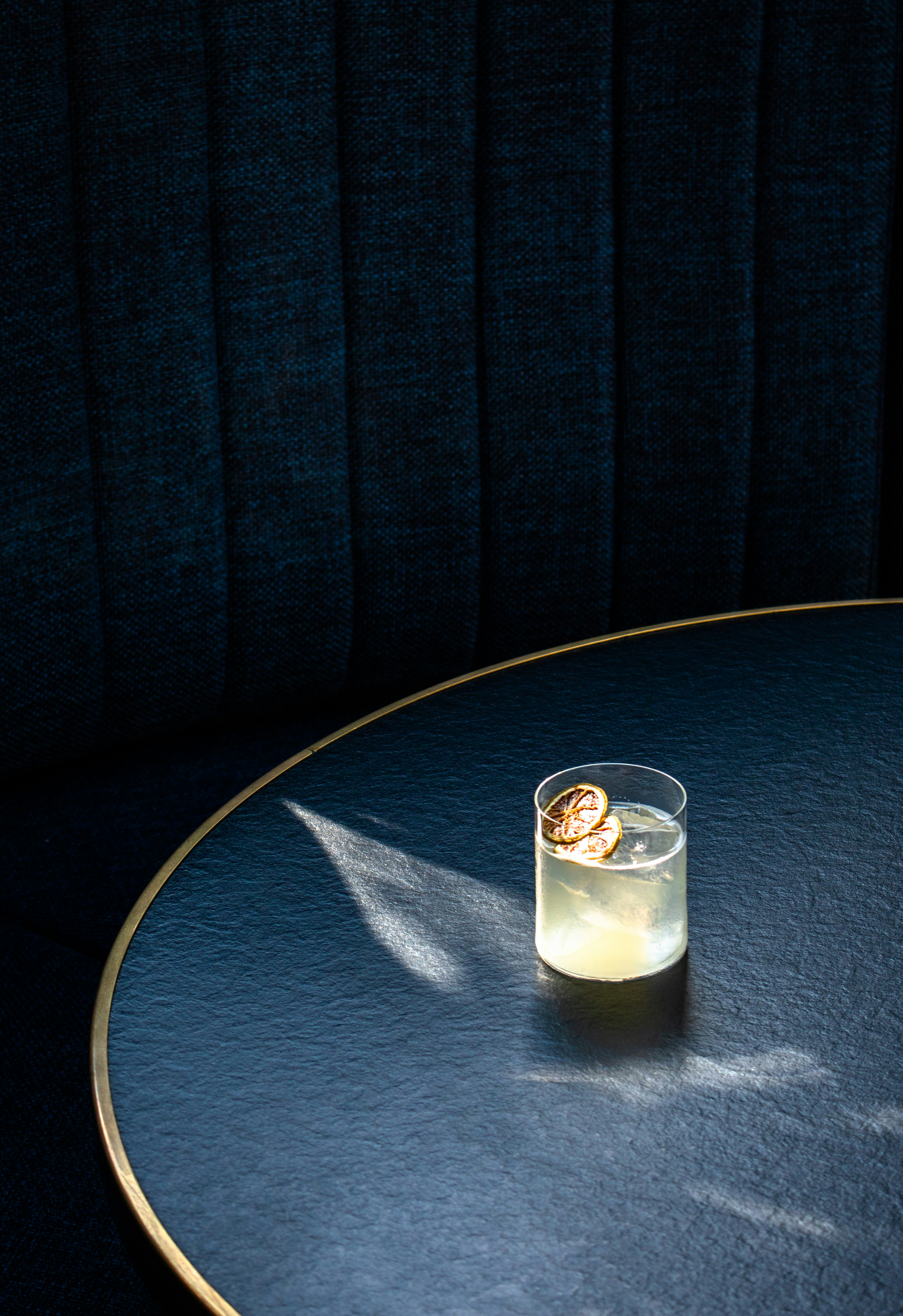 Un verre de cocktail avec des tranches de citron séché sur une table ronde sombre avec une banquette bleu foncé en arrière-plan.