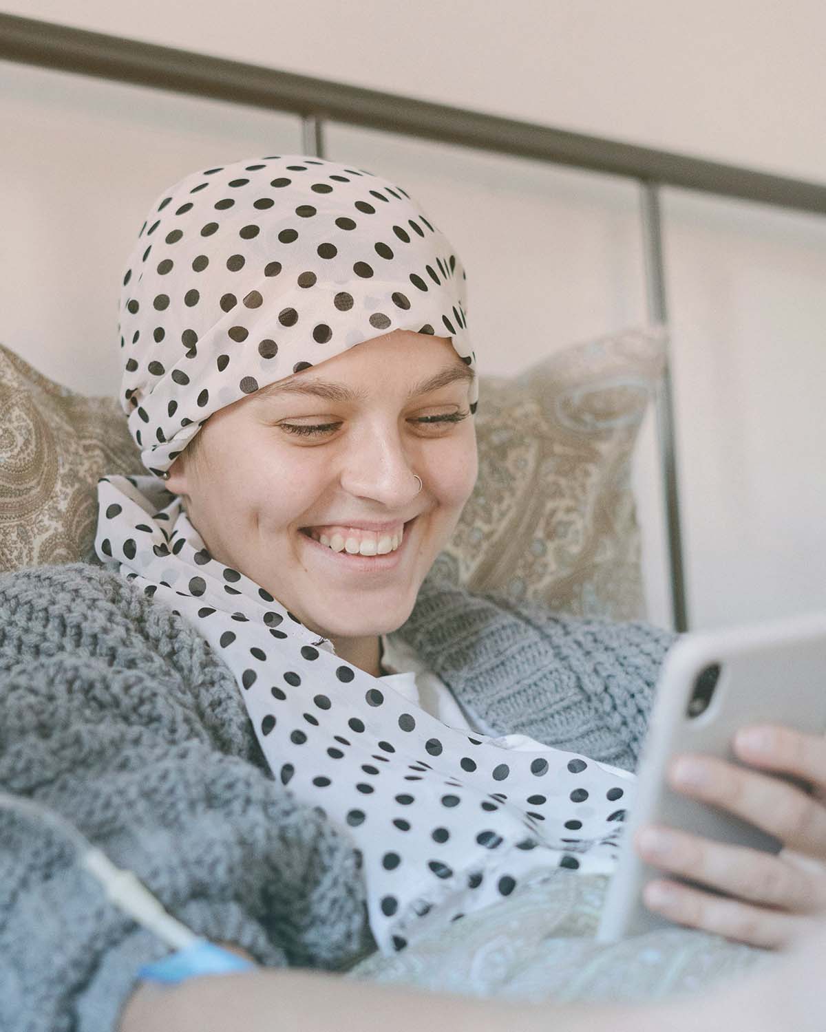 Junge Krebspatientin mit Kopftuch lächelt im Bett und schaut aufs Smartphone – Symbol für Lebensfreude und Zuversicht während der Krebstherapie mit unterstützender Behandlung wie CBD oder THC.