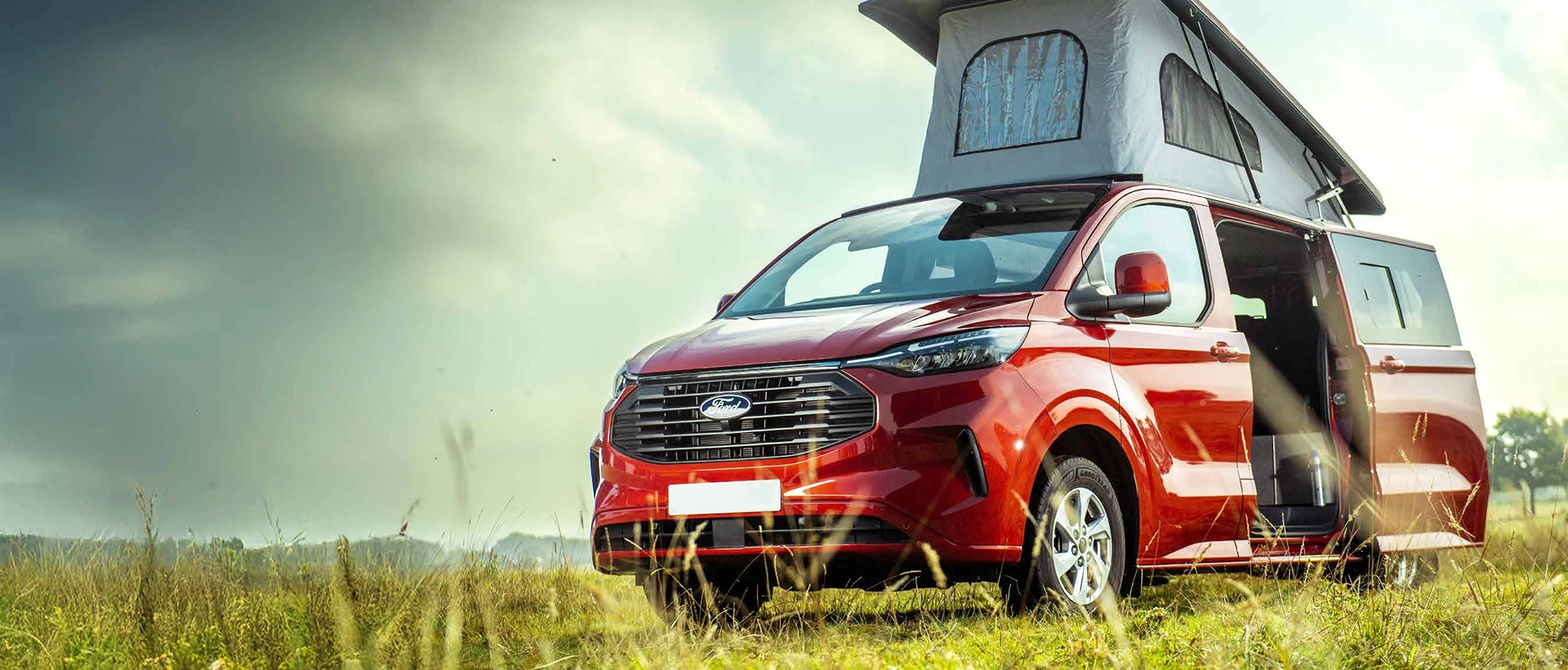 Red Ford camper van with roof tent and open side door parked in a grassy field under a cloudy sky.