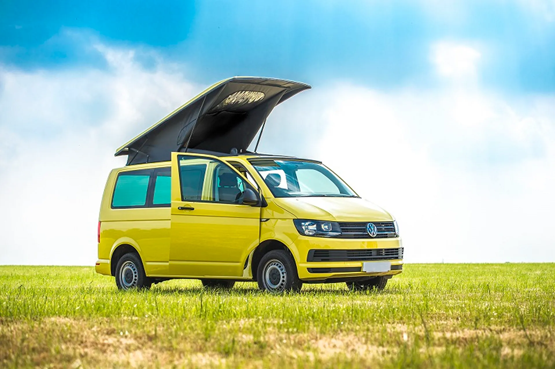 Yellow Volkswagen camper van with pop-up roof and open door parked on green grass under blue sky.