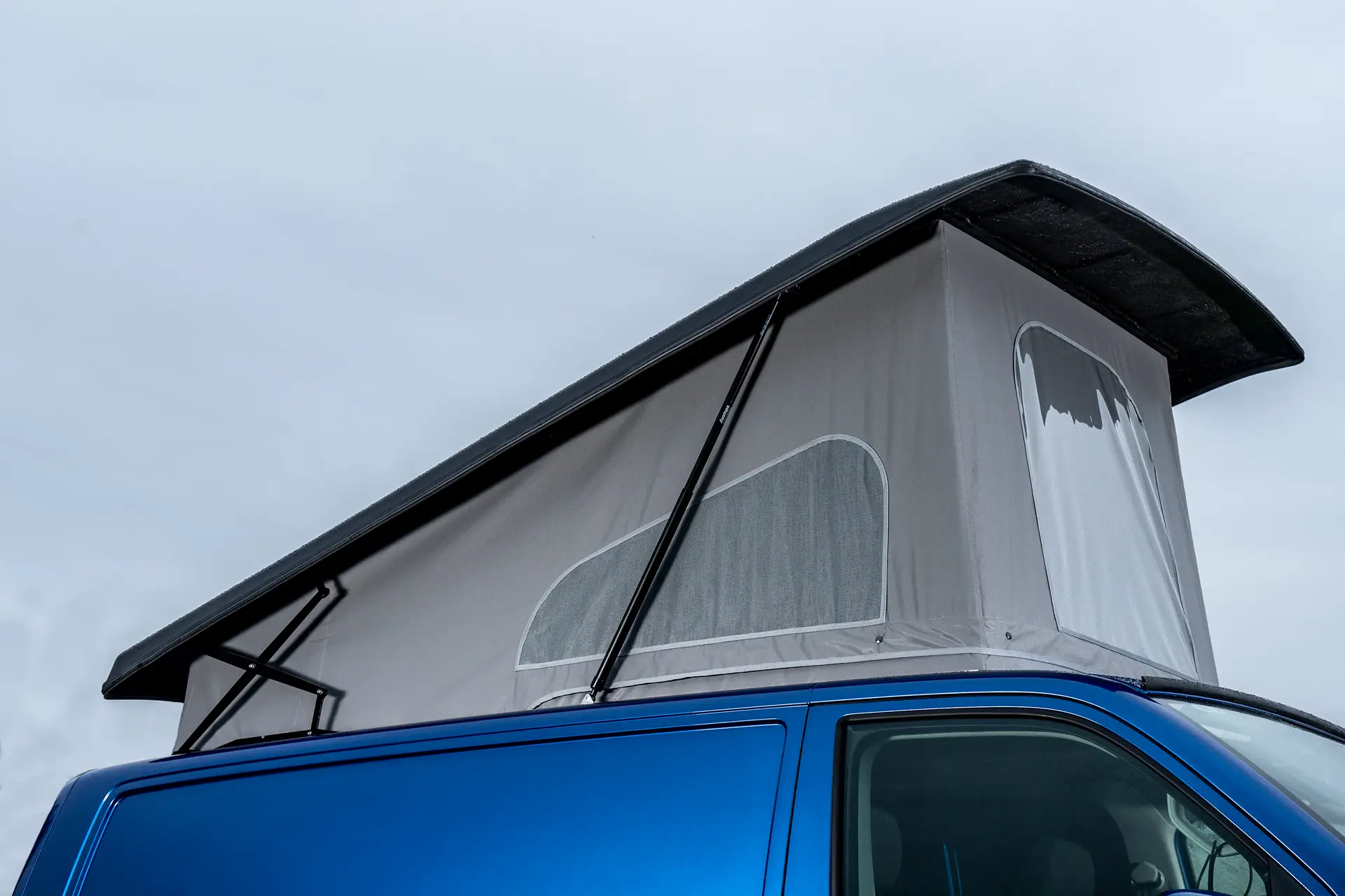 Blue camper van with gray pop-up roof tent extended under cloudy sky.
