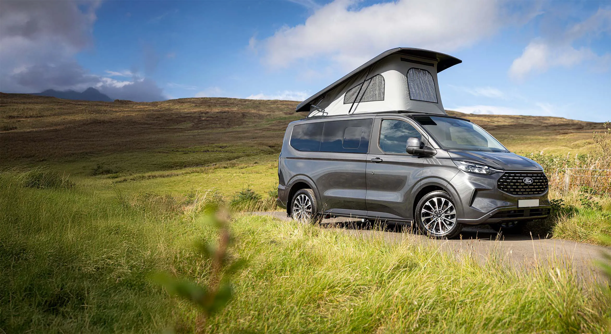 Grey camper van with raised roof parked on a narrow road in a green grassy landscape.