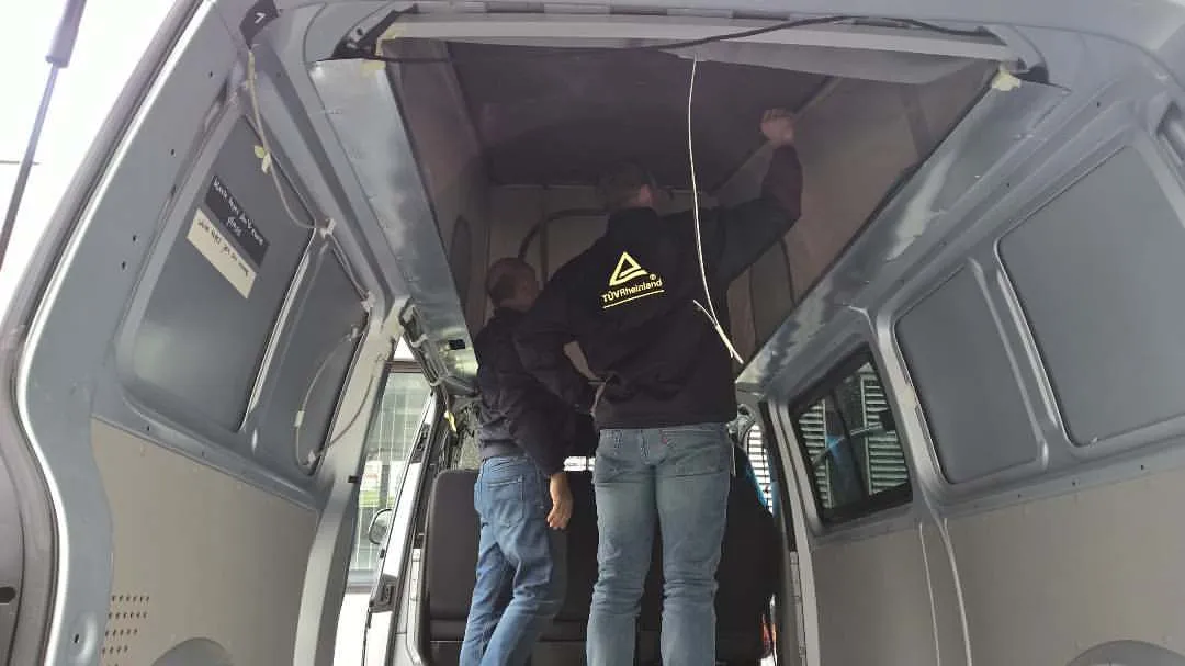 Two men inside a van inspecting or working on the van's ceiling or overhead area.
