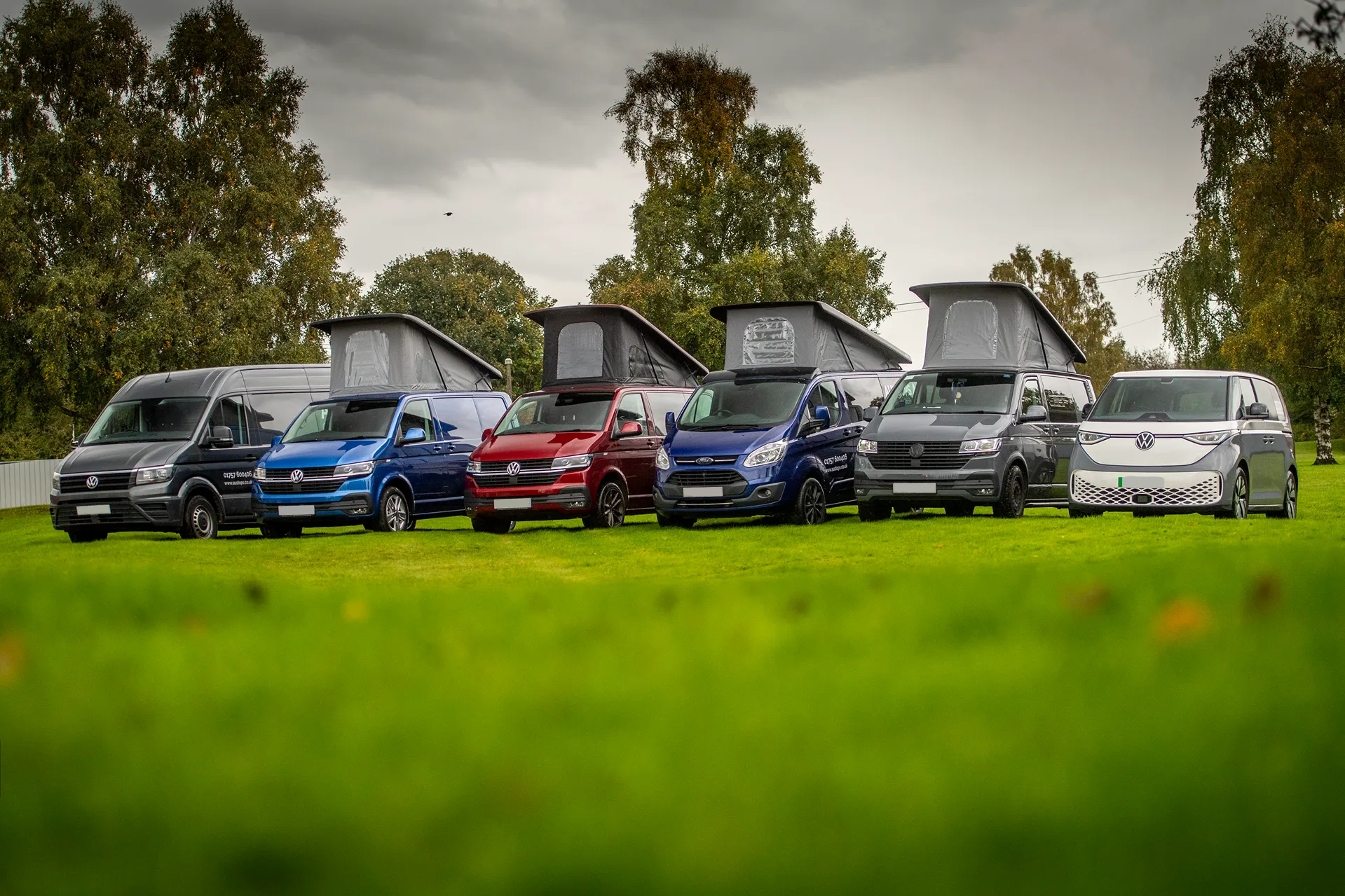 Six camper vans and vans parked on green grass under a cloudy sky with trees in the background.