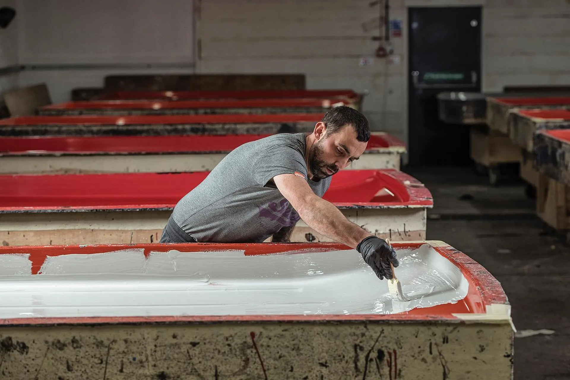 Man wearing gloves applies white paint inside a red frame on a long workbench in a workshop.