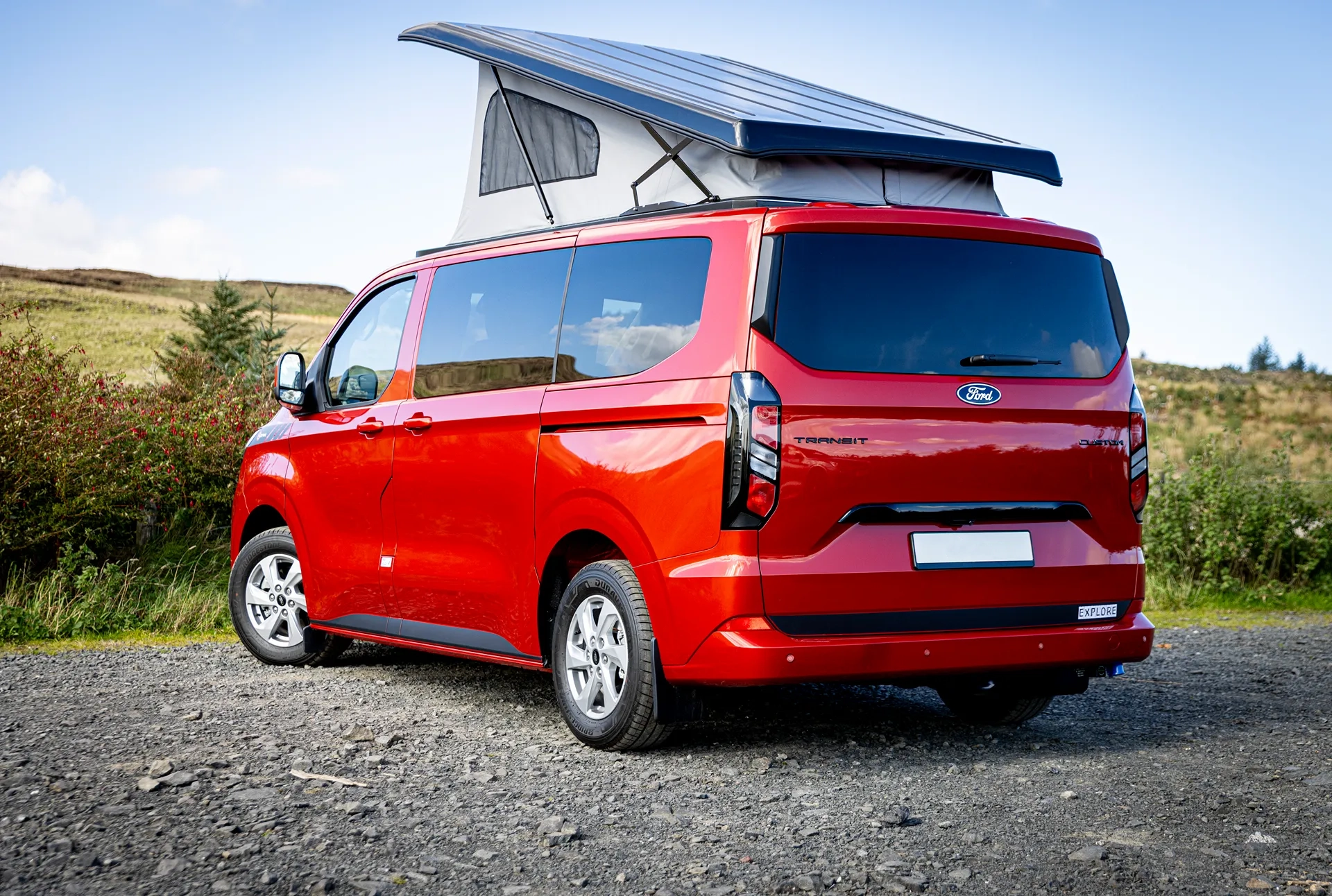 Red Ford Transit camper van with a raised pop-up roof parked on gravel near grass and hills.