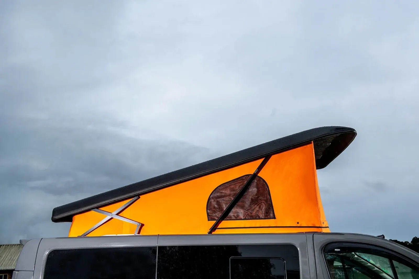 Gray van with orange pop-up roof tent under cloudy sky.