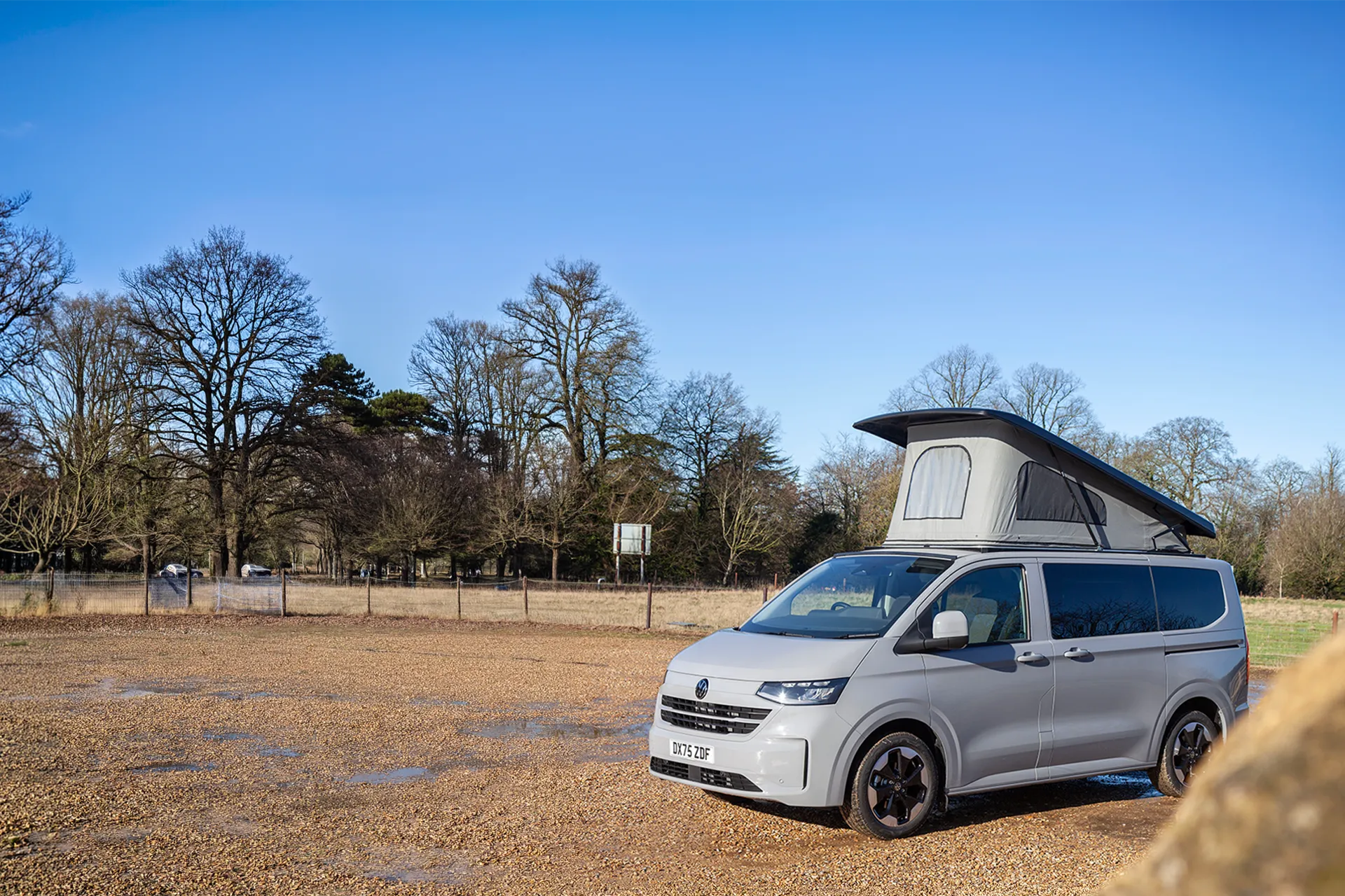 Light gray camper van with raised roof parked on a gravel area near leafless trees under blue sky.