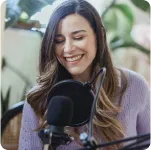 Smiling woman with long hair speaking into a microphone in a casual indoor setting.