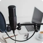 Black microphone with pop filter on a boom arm in front of a laptop and a cup on a white desk.