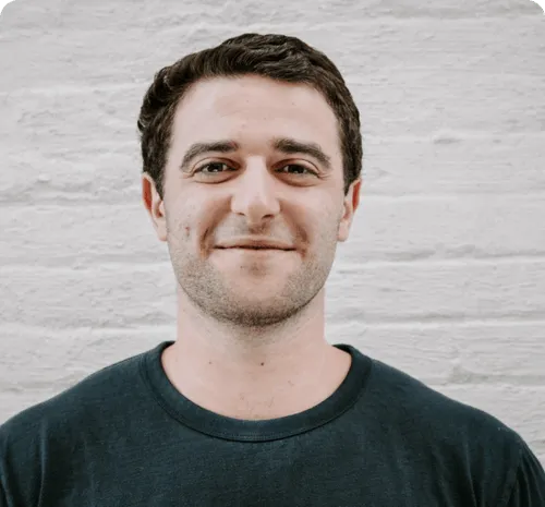 Smiling man with short dark hair wearing a dark crew-neck shirt against a light brick wall background.